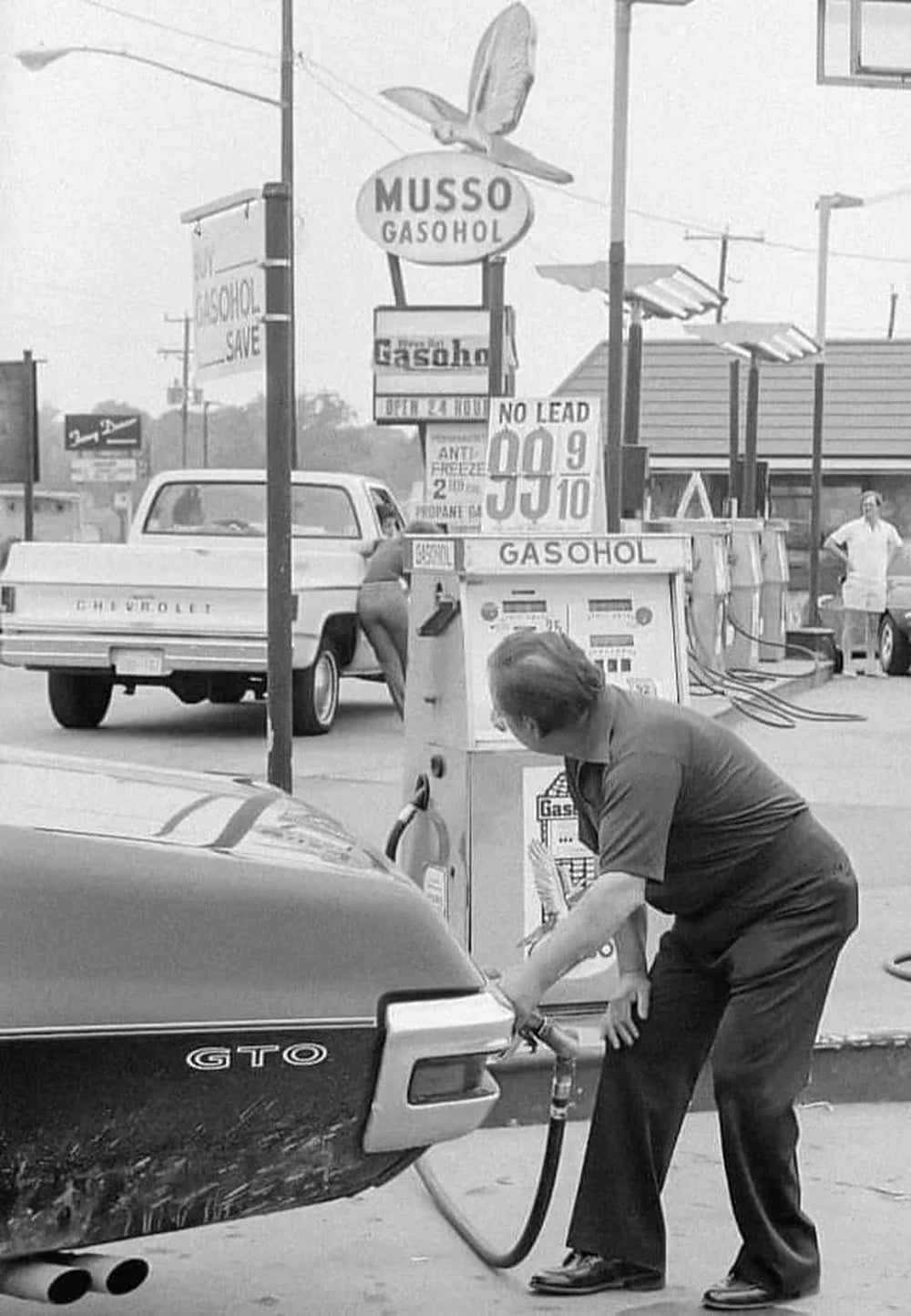 A man in dark clothes pumps gas into a Pontiac GTO at a vintage Musso Gasohol station. A white truck and several people are visible in the background. The image appears to be from the 1970s.
