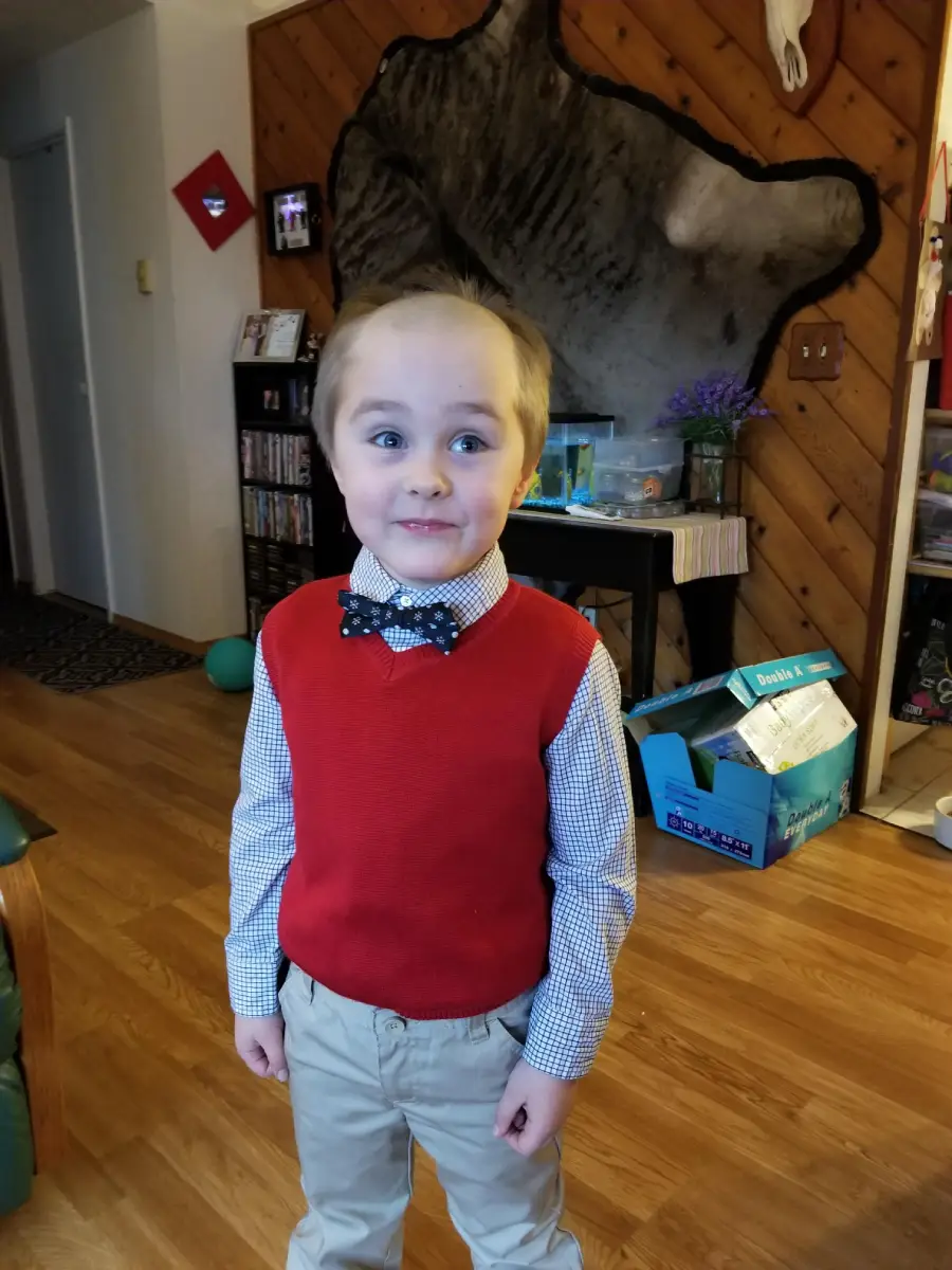A young child in a red sweater vest, checkered shirt, and bow tie stands indoors on a wood floor, smiling with wide eyes. A large animal pelt hangs on the wood-paneled wall behind him.