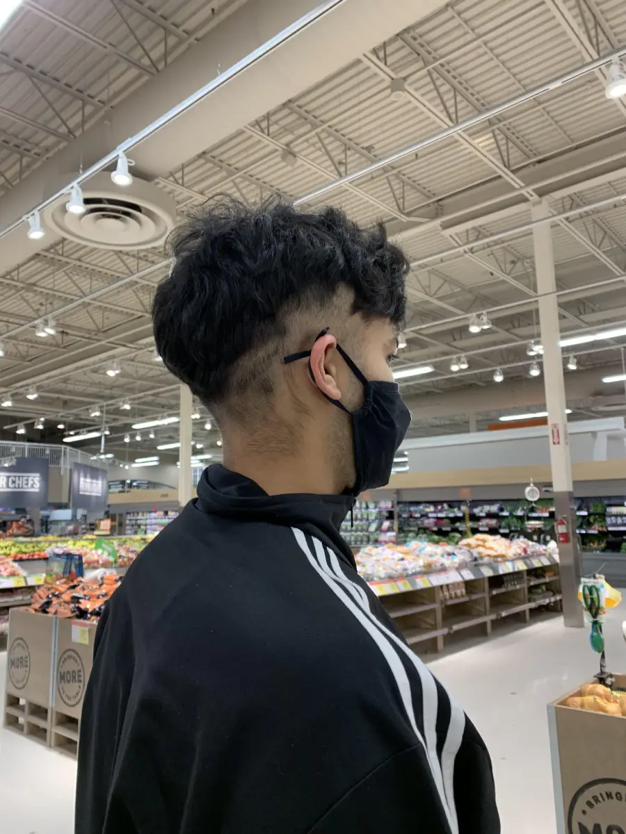 A person with curly dark hair and a black mask stands in a grocery store produce section, wearing a black jacket with white stripes on the sleeves.