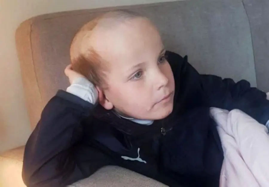 A young child with light hair sits on a gray couch, resting their head on one hand and looking to the side with a calm expression. They are wearing a dark jacket with a white logo.