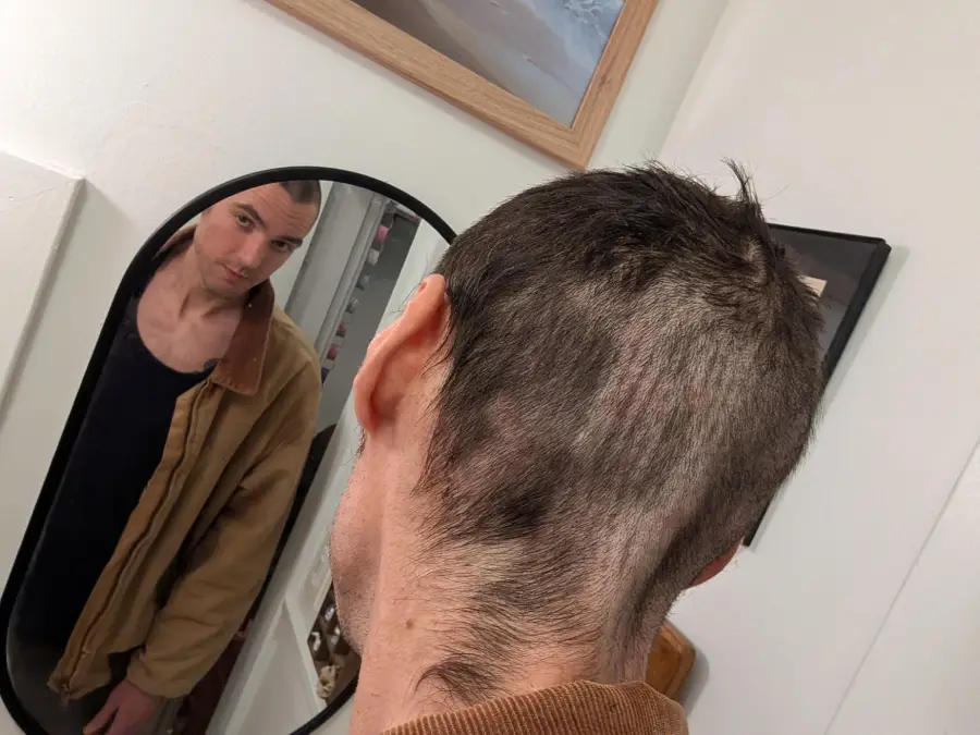 A person looks into a mirror, showing the back of their head with an uneven, partially shaved haircut. They wear a brown jacket and dark shirt, standing in a white room with framed pictures on the walls.