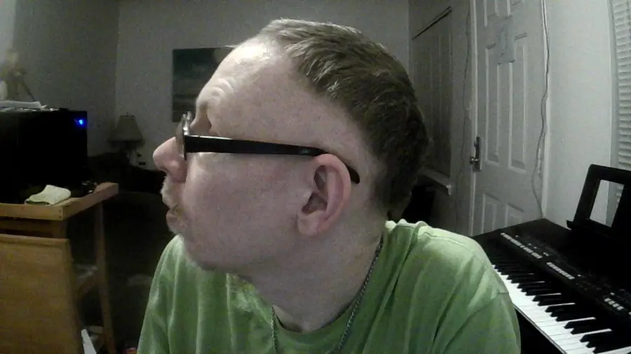 A person with short hair and glasses looks to the left in a dimly lit room. A keyboard, window blinds, and a door are visible in the background. The person is wearing a green shirt and a necklace.