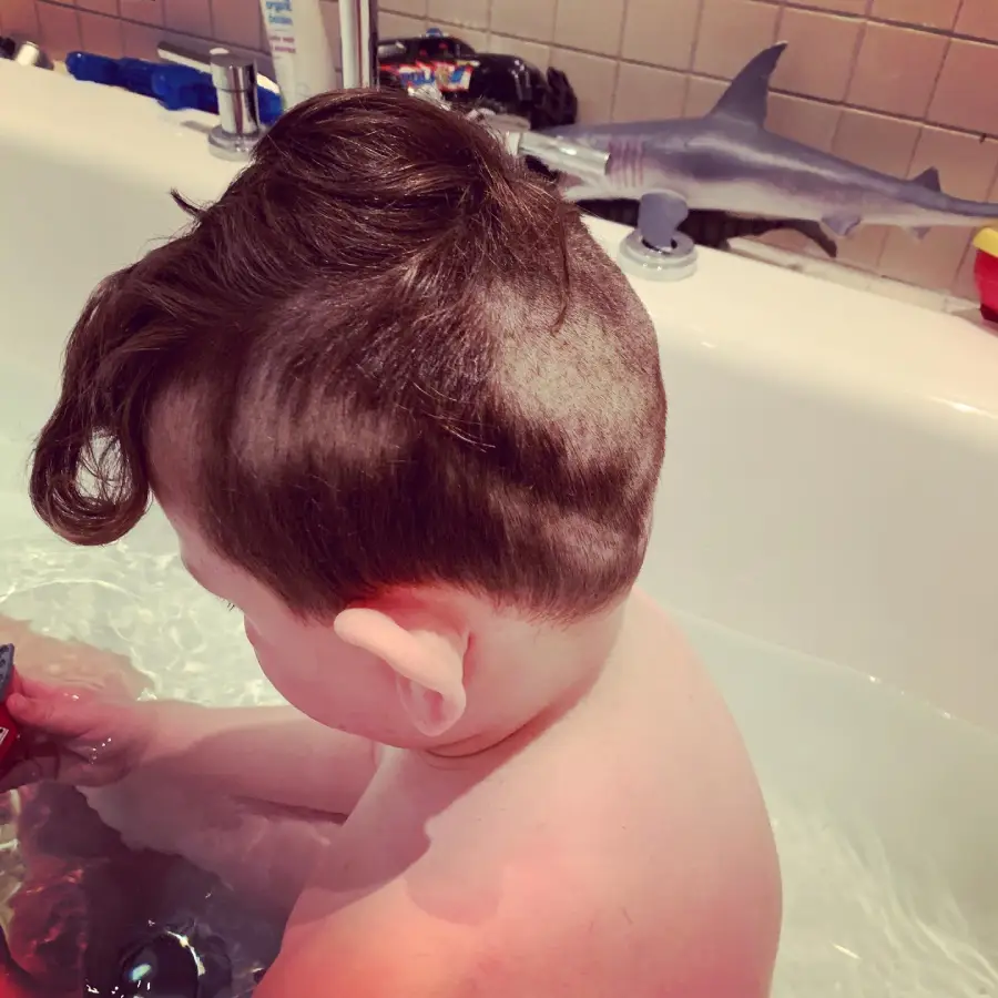 A child with a partially shaved haircut sits in a bathtub, holding a toy. A toy shark and other bath items are visible on the edge of the tub in the background.