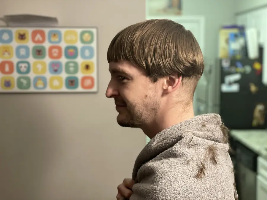 A man with light brown hair and a bowl haircut smiles slightly in a kitchen. He wears a gray towel around his shoulders, and there is hair on the towel. Colorful icons are displayed on the wall behind him.