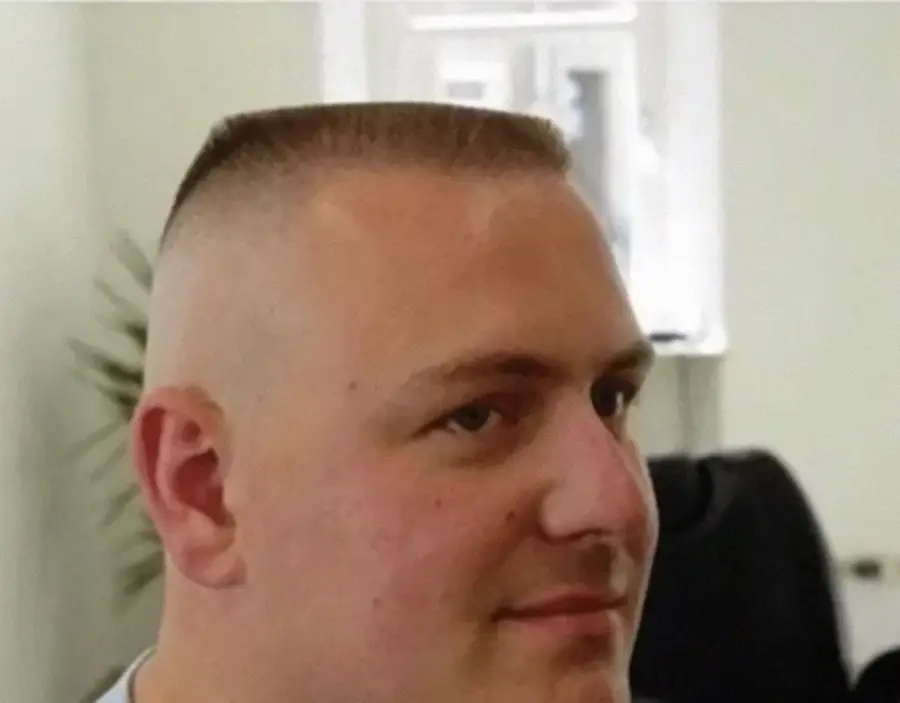 A person with a flat-top haircut, where the hair on top is cut flat and very short, while the sides are shaved closely. The background is an indoor setting with blurred details.