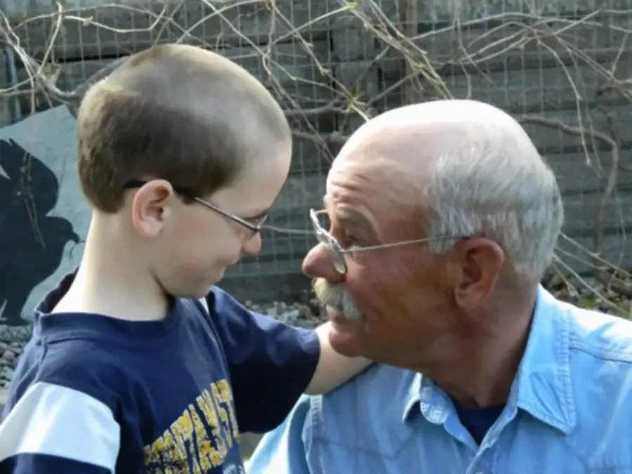 An older man and a young boy smile warmly at each other outdoors, with the man’s arm around the boy’s shoulder. They appear to share a close, affectionate moment.