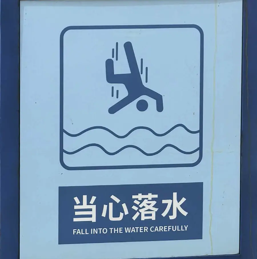 A sign shows a person falling into water above the text “FALL INTO THE WATER CAREFULLY” and Chinese characters, warning people to be cautious near the water.