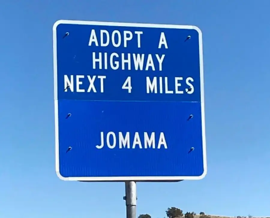 Blue road sign reads: “Adopt A Highway Next 4 Miles JOMAMA” against a clear blue sky. Trees are faintly visible at the bottom of the image.
