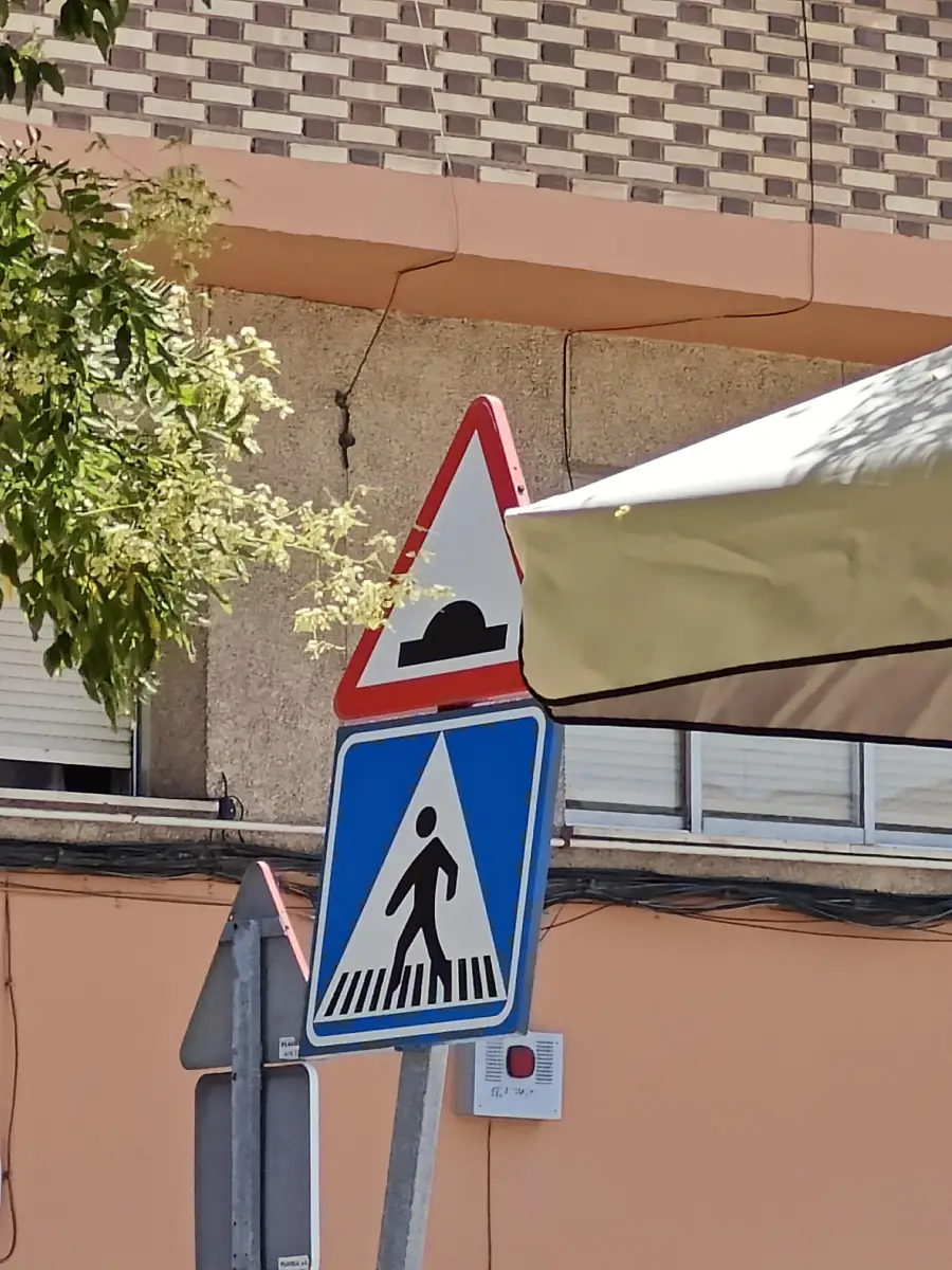 A triangular bump warning sign is above a blue pedestrian crossing sign. Both are partly shaded by a tree and a beige umbrella. A beige building with windows is in the background.