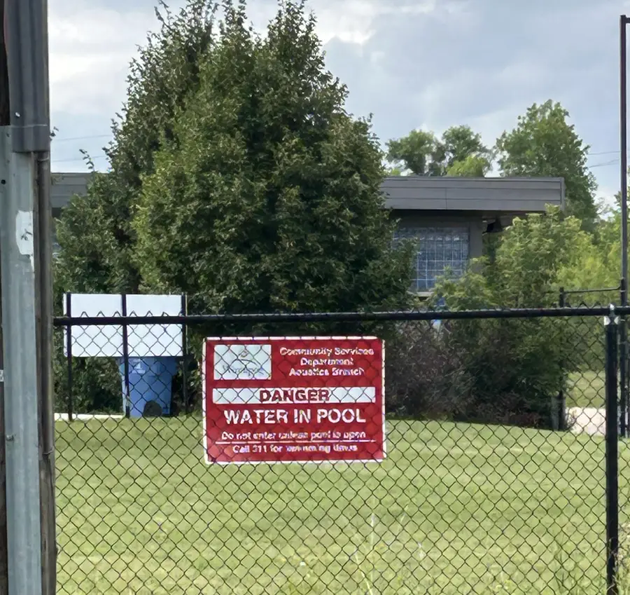 A red sign on a chain-link fence warns “Danger: Water in Pool” in front of a grassy area, large trees, and a building in the background under a cloudy sky.