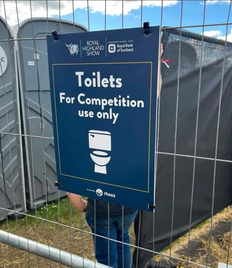 A sign on a fence reads, “Toilets For Competition use only,” with a toilet icon. Portable toilets are visible behind the fence at an outdoor event.