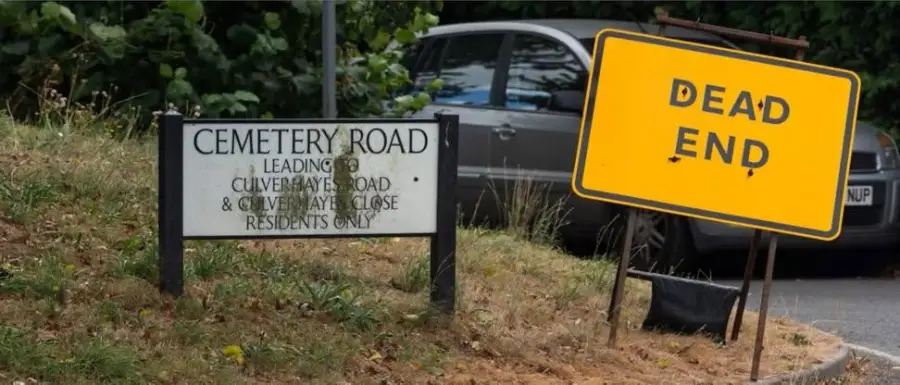 A sign reading “Cemetery Road” stands next to a yellow “Dead End” sign beside a parked car and greenery in the background.