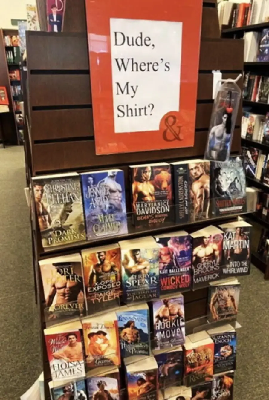 A bookstore display features shirtless men on romance novel covers. A sign above reads “Dude, Where’s My Shirt?” in large text. Books are arranged neatly on multiple shelves below the sign.