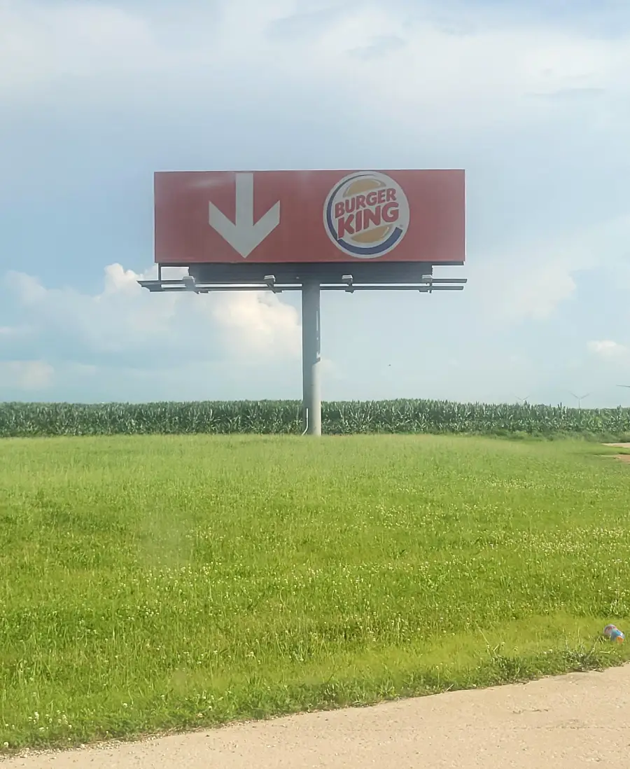A roadside billboard shows a large white arrow pointing down next to the Burger King logo, set against a grassy field and blue sky.