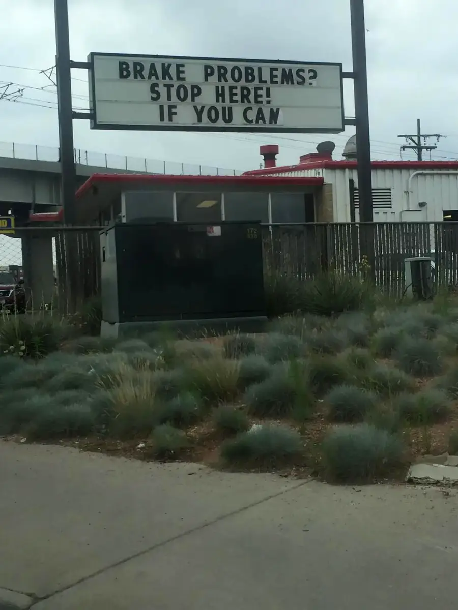 A sign reads, “Brake problems? Stop here! If you can,” above a patch of grass by the road, with a building and utility box in the background.