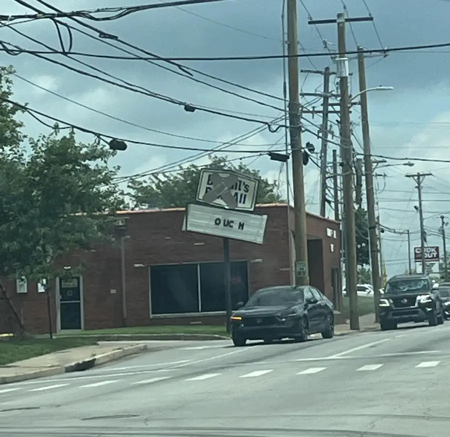 A brick building with a damaged tilted sign on top reads “OUCH” in large letters. Two cars are driving past on the street under cloudy skies.