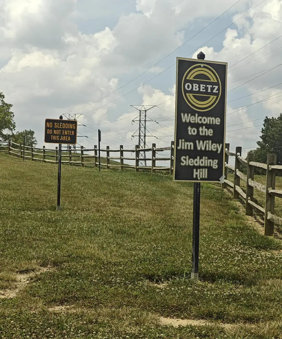 A grassy hill with two signs; one says “Welcome to the Jim Wiley Sledding Hill,” and the other says “No Sledding. Do Not Enter This Area,” with a wooden fence and power lines in the background.