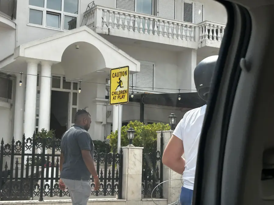 A “Caution: Children at Play” sign is posted outside a white building with columns. A man walks nearby, and a person in a helmet is partially visible in the foreground, seen from a car window.