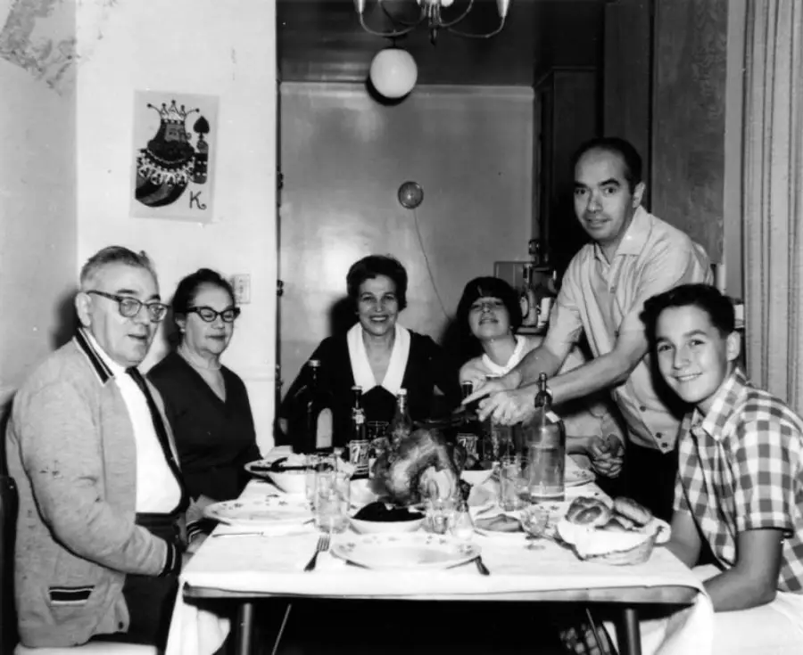 Six people sit around a dining table set for a meal, smiling at the camera. There is food on the table, including a roast. The room is decorated with balloons and a “Happy Birthday” sign.