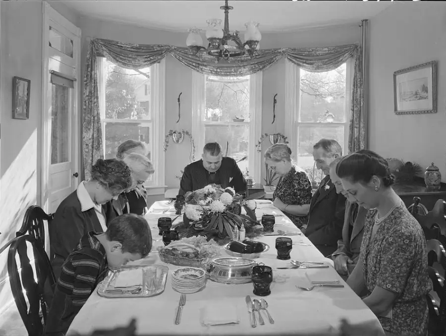 Eight people sit around a dining table with food, heads bowed in prayer. The scene appears to be from the mid-20th century, with sunlight streaming through large windows behind them.