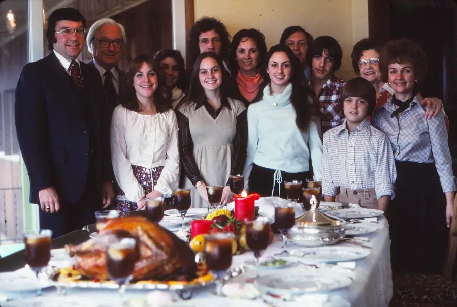 A family poses and smiles together behind a dining table set for a meal, with a roast turkey, candles, and drinks visible, suggesting a holiday or special gathering.