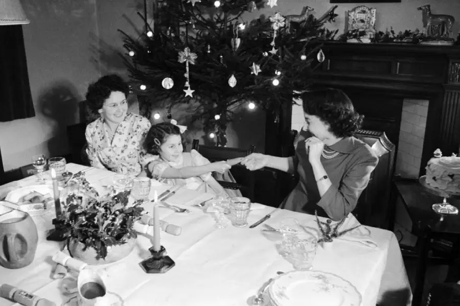 Three people sit at a decorated table, pulling a Christmas cracker. A Christmas tree with lights and ornaments stands behind them, creating a festive atmosphere.