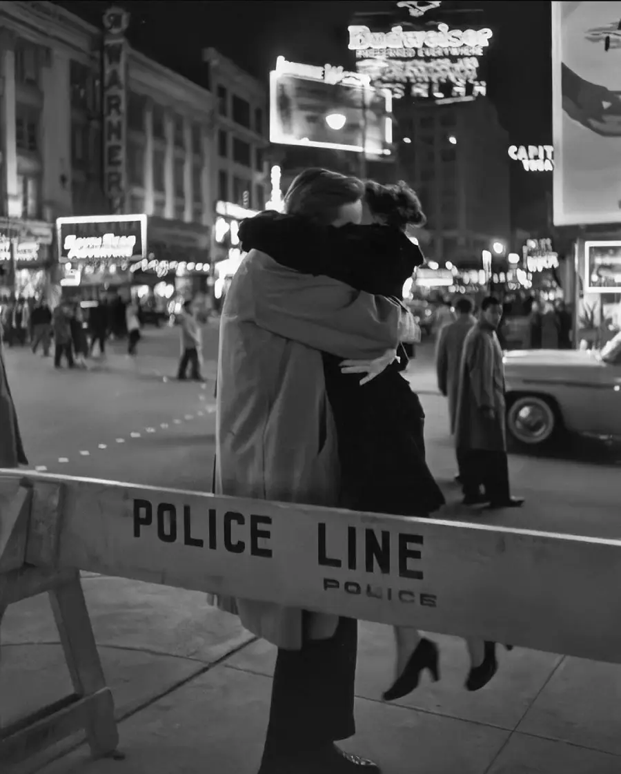 A couple embraces passionately on a city street at night behind a police line barrier, with neon signs and vintage cars in the background.
