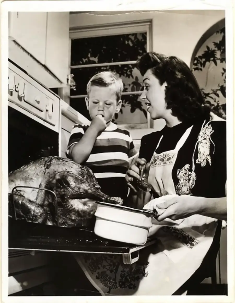 A woman in an apron bastes a turkey in the oven while a young boy beside her holds his nose, looking displeased. The photo is black and white, set in a vintage kitchen.