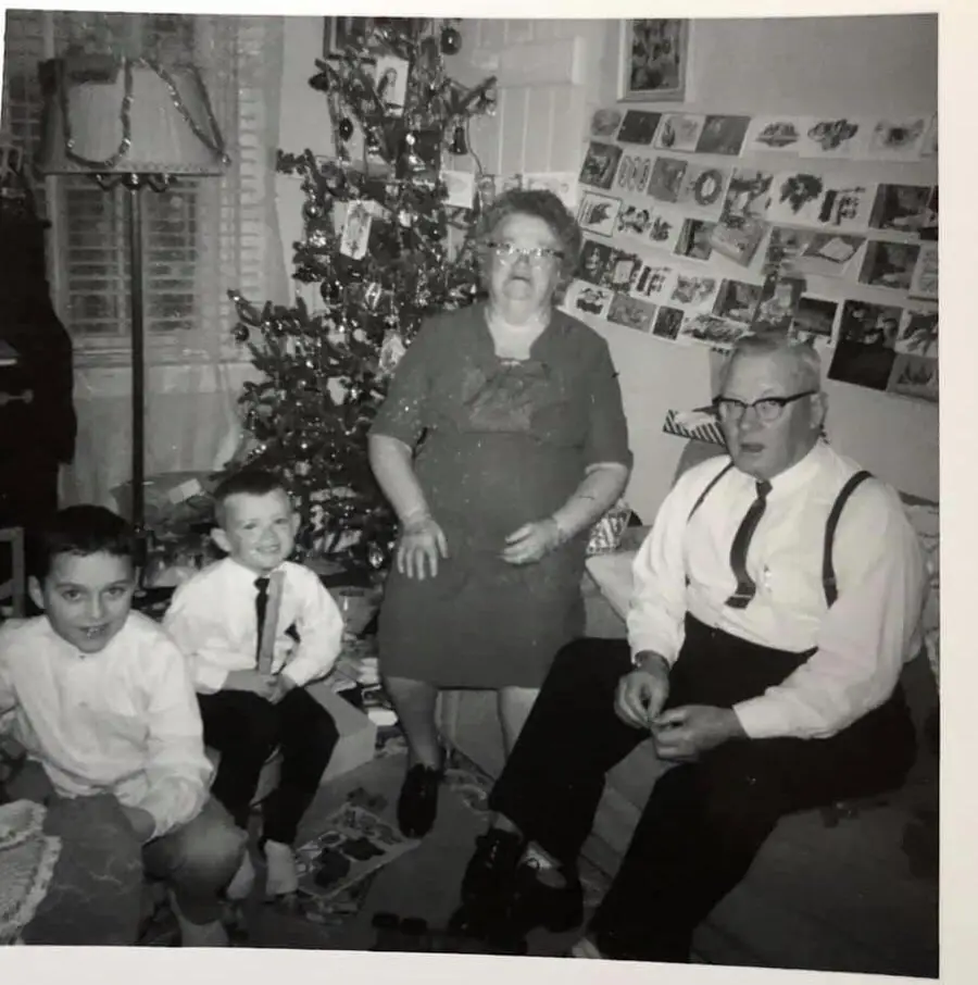 Black and white photo of a family in front of a decorated Christmas tree; two boys in white shirts, an older woman in a dress, and an older man with glasses and suspenders. Holiday cards are displayed on the wall.