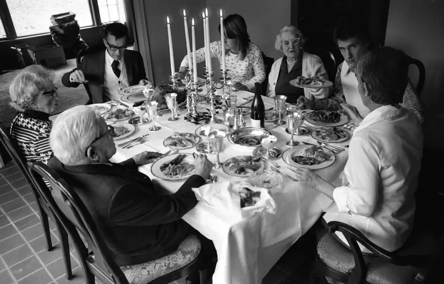 Seven people sit around a dining table eating a meal together. The table is set with plates of food, tall candles, and silverware. The scene is indoors, and the photo is in black and white.
