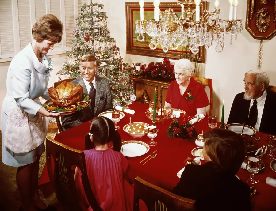 A woman serves roast turkey at a holiday dinner table with six people, including children and elderly, decorated with a Christmas tree, candles, and festive ornaments.