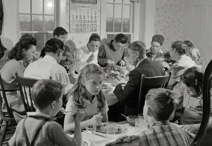 A large family sits around a dining table sharing a meal. Children and adults are engaged in eating and conversation. Sunlight streams through the windows behind them. A calendar hangs on the wall.