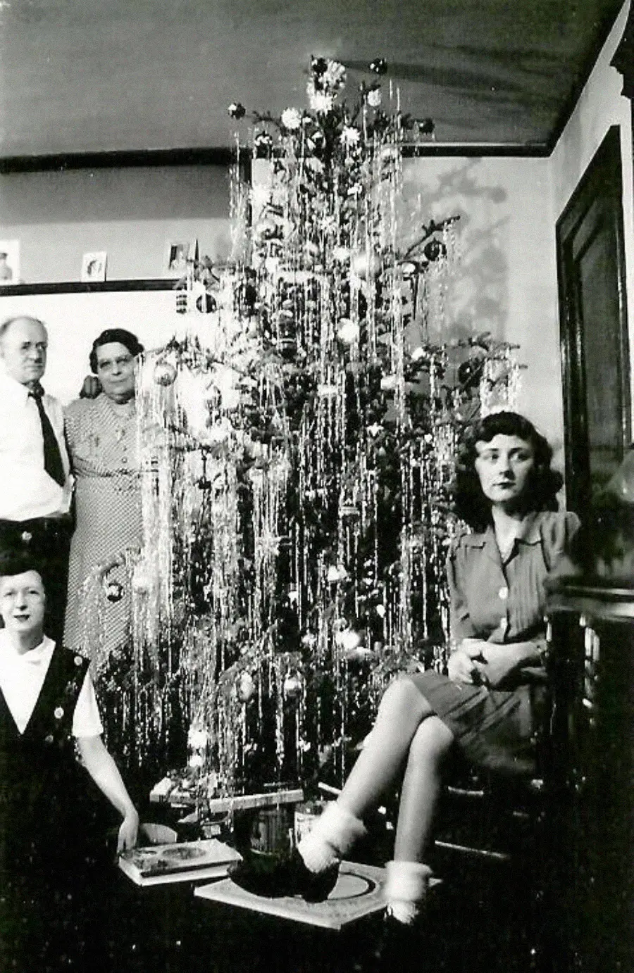 Four people pose beside a tall, tinsel-covered Christmas tree in a living room. Two adults stand, while a girl sits on stairs and a boy kneels on the floor. The room is decorated for the holiday.