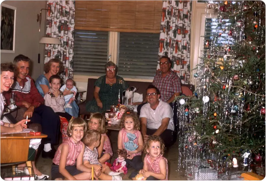A group of adults and children gather in a living room by a decorated Christmas tree, smiling for a photo. Some children hold dolls and toys, while wrapped gifts are visible on the table and floor.