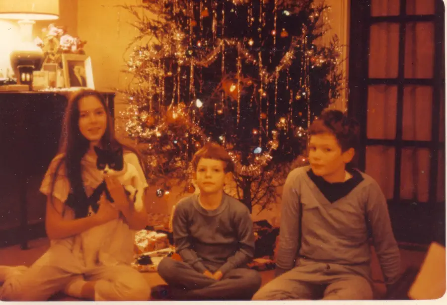 Three children sit in front of a decorated Christmas tree. The girl on the left holds a black and white cat. Holiday decorations and wrapped gifts are visible under the tree. Warm lighting fills the cozy room.