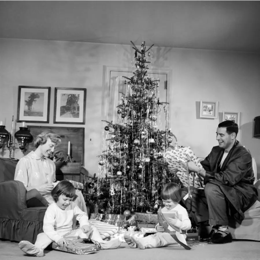 A family of four sits by a decorated Christmas tree, opening presents. The parents watch happily as two young children unwrap gifts on the floor. The scene is festive and cozy, with ornaments and tinsel on the tree.