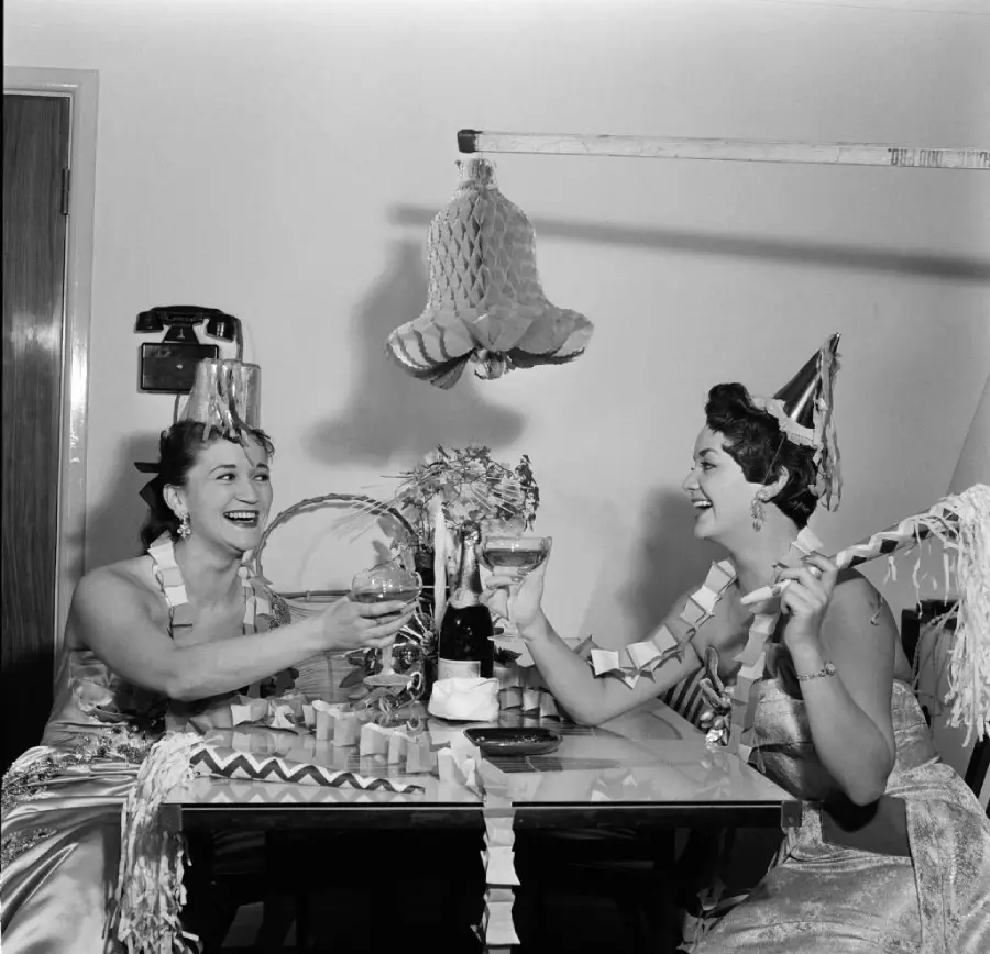 Two women in party hats sit at a decorated table, clinking glasses and smiling, surrounded by streamers and festive decorations, celebrating at what appears to be a New Year’s party.