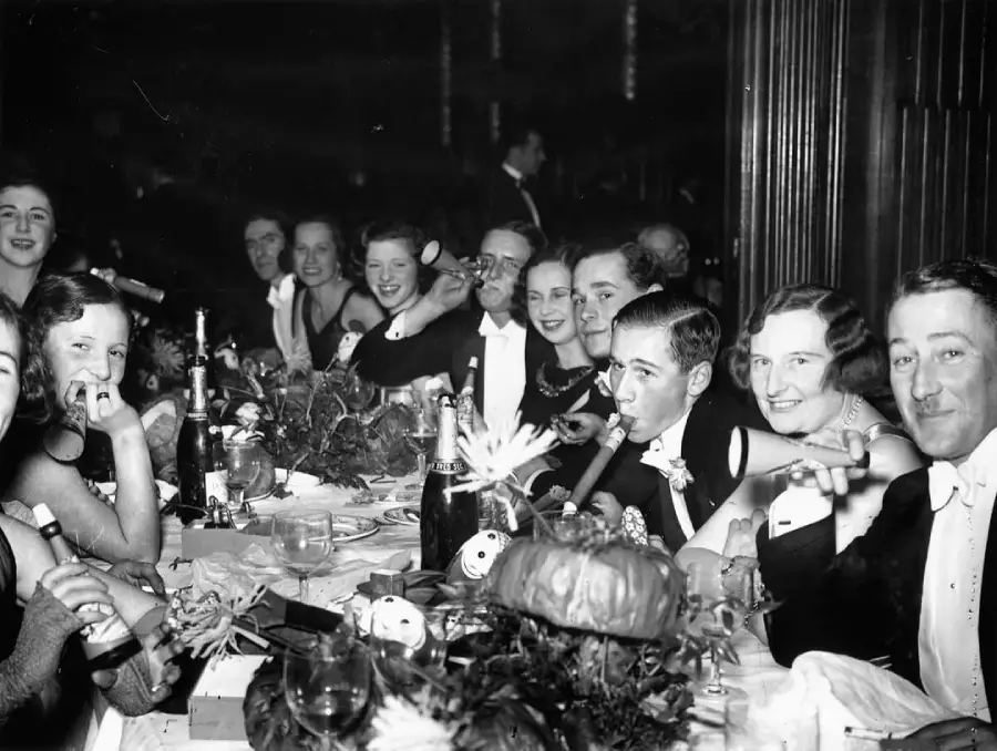 A group of people dressed formally sit around a banquet table, smiling and enjoying drinks at a lively party. Bottles, glasses, and table decorations are visible in the foreground.