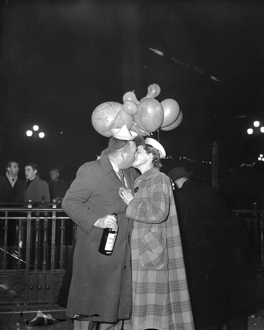A couple wearing party hats kisses on a city street at night. One holds balloons and a bottle, while people and streetlights appear in the background.