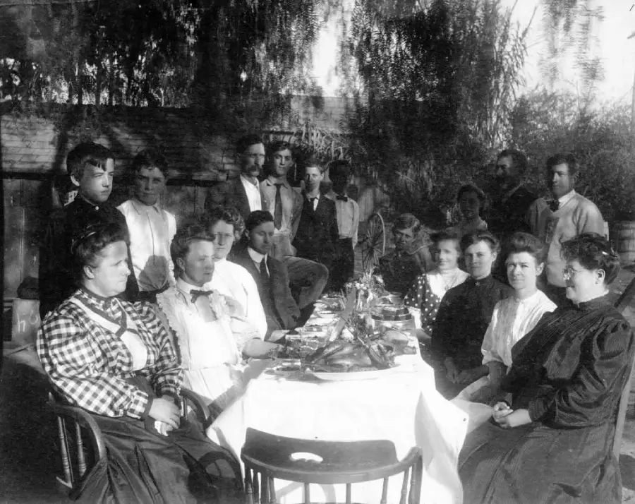 A group of men and women sit and stand around a long outdoor table set with food, posing for a black-and-white photo under trees, dressed in early 20th-century clothing.