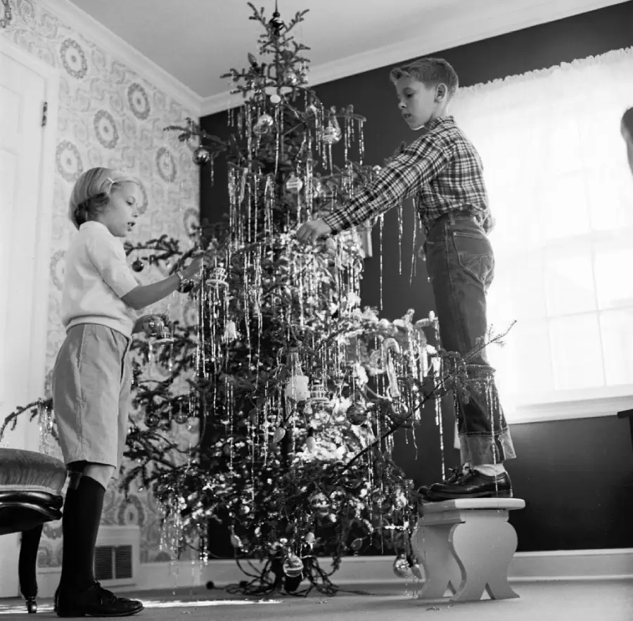 Two children decorate a Christmas tree with tinsel in a living room. One child stands on a stool to reach higher branches, while the other stands nearby, both focused on the tree.