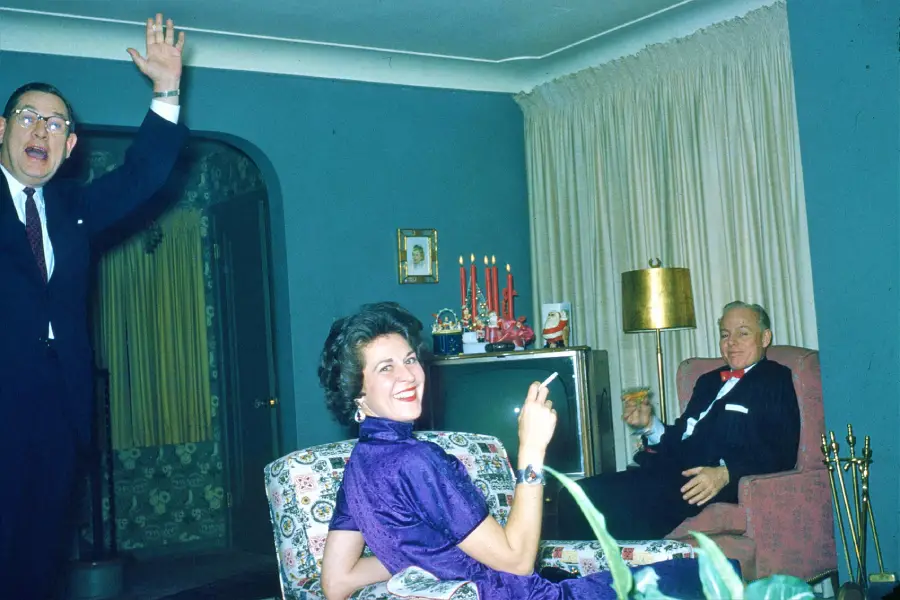 A woman in a purple dress smiles while holding a cigarette, a man waves enthusiastically on the left, and another man sits in a chair holding a drink in a cozy, mid-century living room.