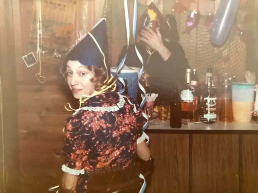A person in a festive outfit and party hat sits at a table, looking back at the camera. Bottles and drinks are on the table, and another person in a hat is partly visible in the background.
