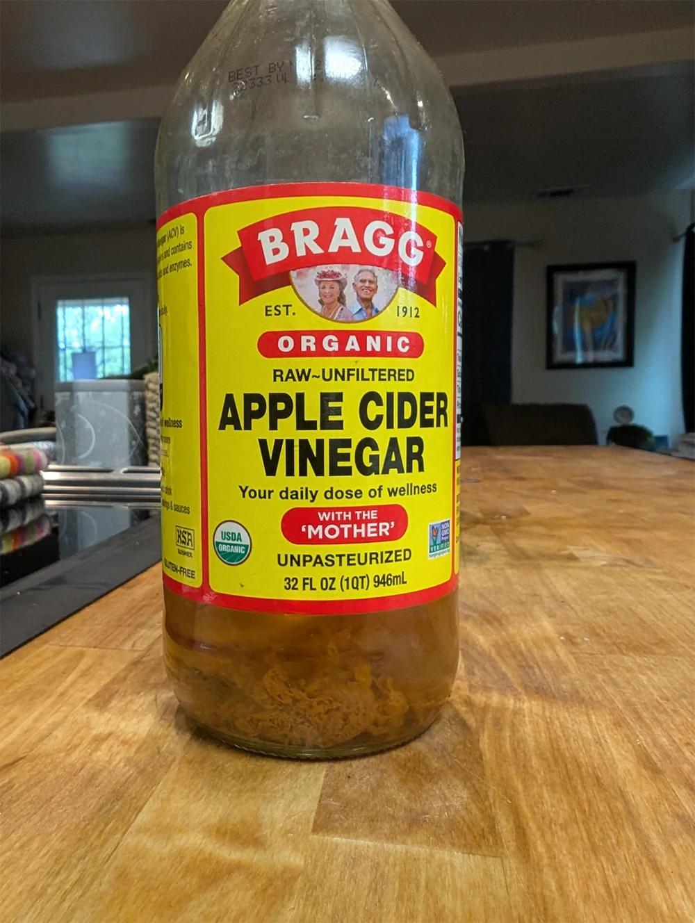A bottle of Bragg Organic Raw Unfiltered Apple Cider Vinegar with “With the Mother” on the label, sitting on a kitchen counter. The bottle is partially filled.