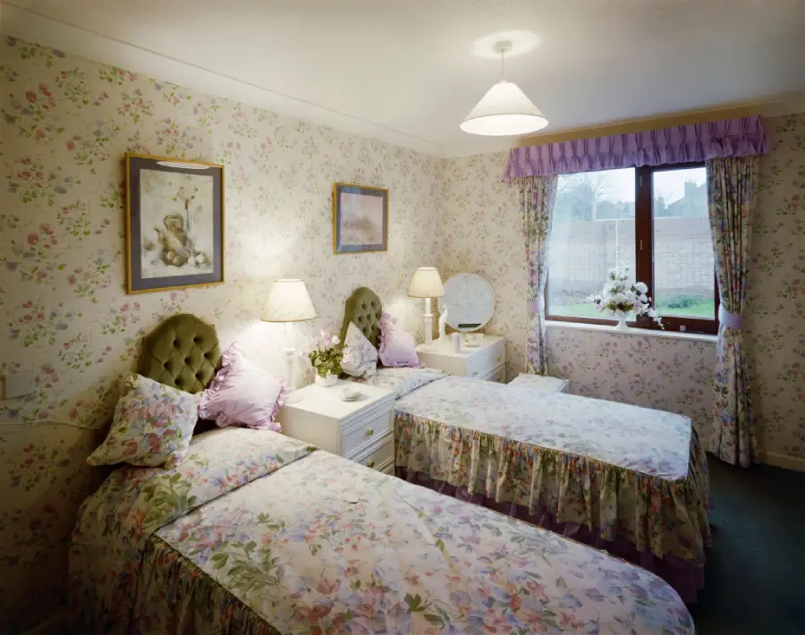 A bedroom with two twin beds, floral bedspreads, matching wallpaper, and curtains. Each bed has a nightstand with a lamp. A window lets in daylight. Framed prints hang on the wall above the beds.
