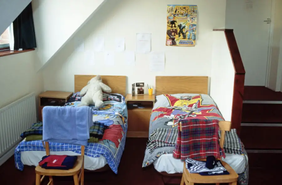 Two single beds with superhero bedding are side by side in a tidy room. A teddy bear is on one bed. Chairs with folded clothes are at the bed ends. A poster and papers are on the wall above the beds.