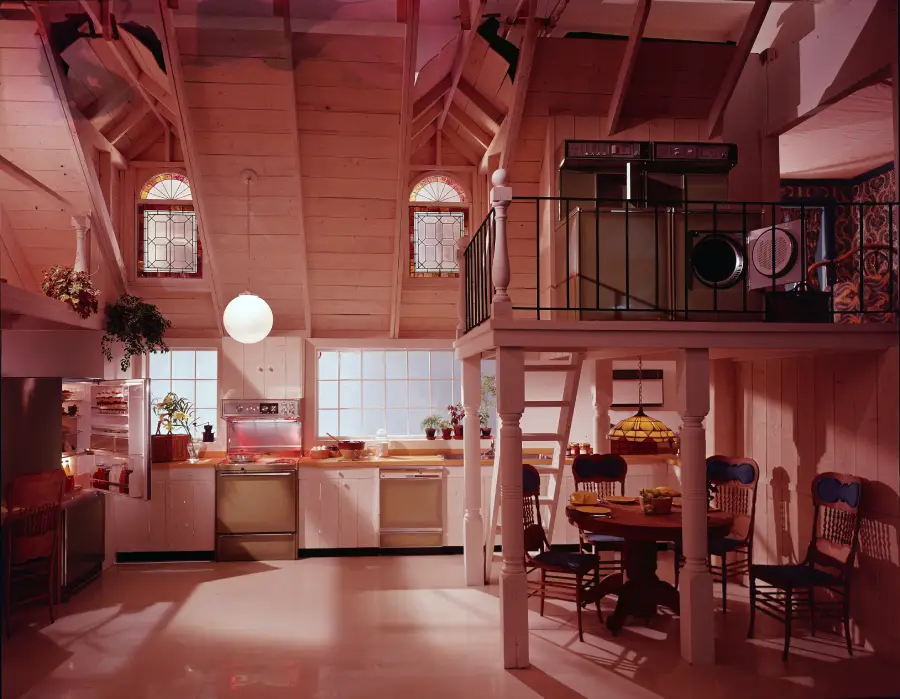 A cozy kitchen with vaulted wooden ceilings, large windows, hanging lights, plants, and a loft above a dining area with a round table and wooden chairs. Stained glass windows add color and charm.
