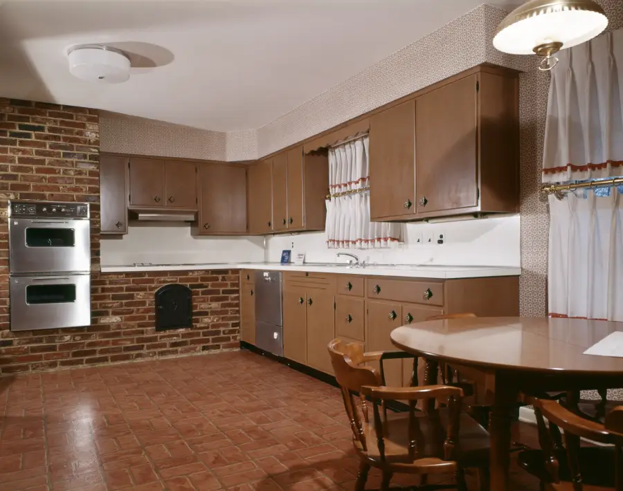 A vintage kitchen with brown cabinets, brick wall, double oven, dishwasher, round wooden table, and chairs. The floor is tiled and windows have frilly white curtains with red trim.
