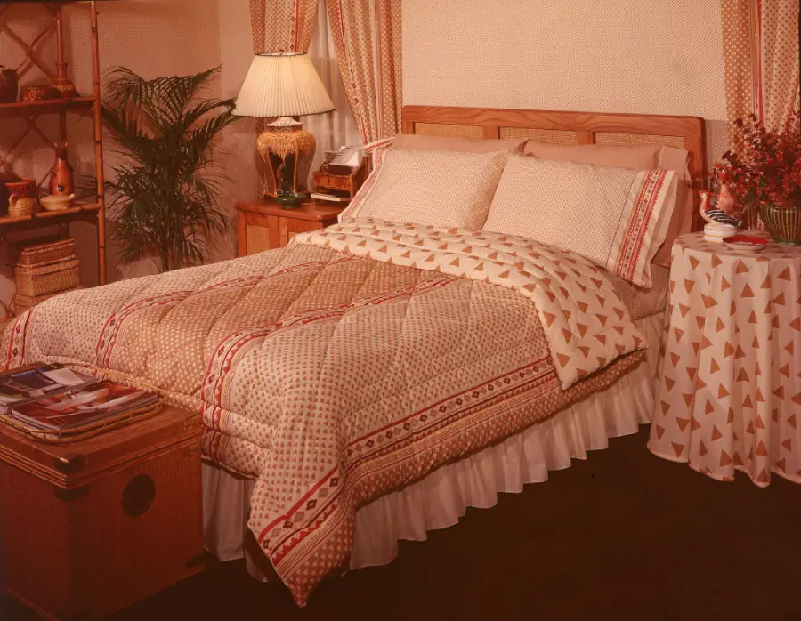 A neatly made bed with a patterned comforter, pillows, and a wooden headboard sits beside a nightstand with a lamp, plants, and a table covered with a matching cloth in a cozy, warmly lit bedroom.