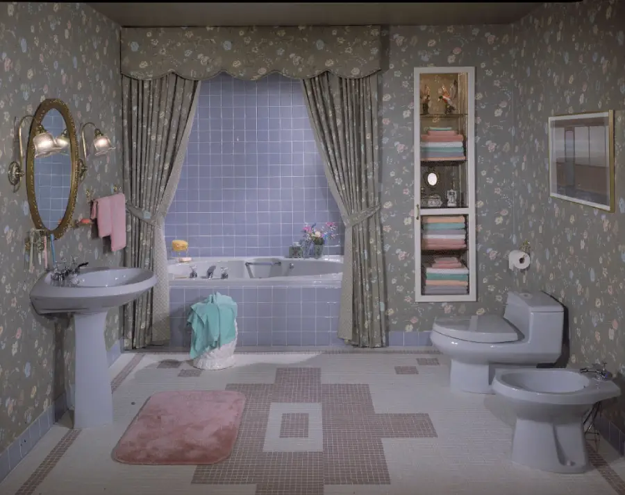 A vintage bathroom with floral wallpaper, pedestal sink, bidet, toilet, bathtub, pink rug, and shelves with neatly folded towels. Curtains frame a window above the tub.
